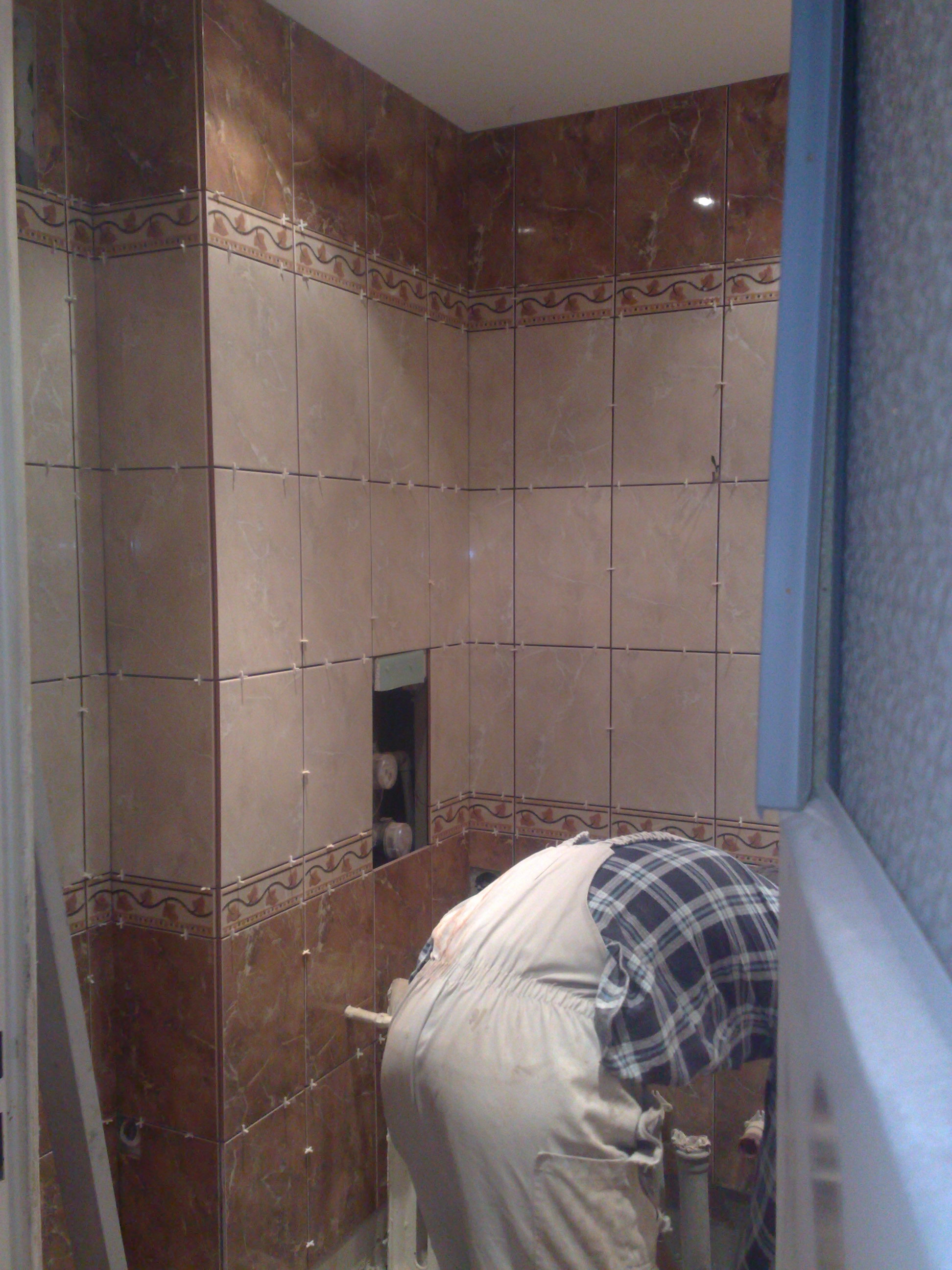 Glazurnik w trakcie układania brązowo-beżowych płytek z dekoracyjnym pasem w łazience, widoczne krzyżyki dystansowe i otwór na rury.