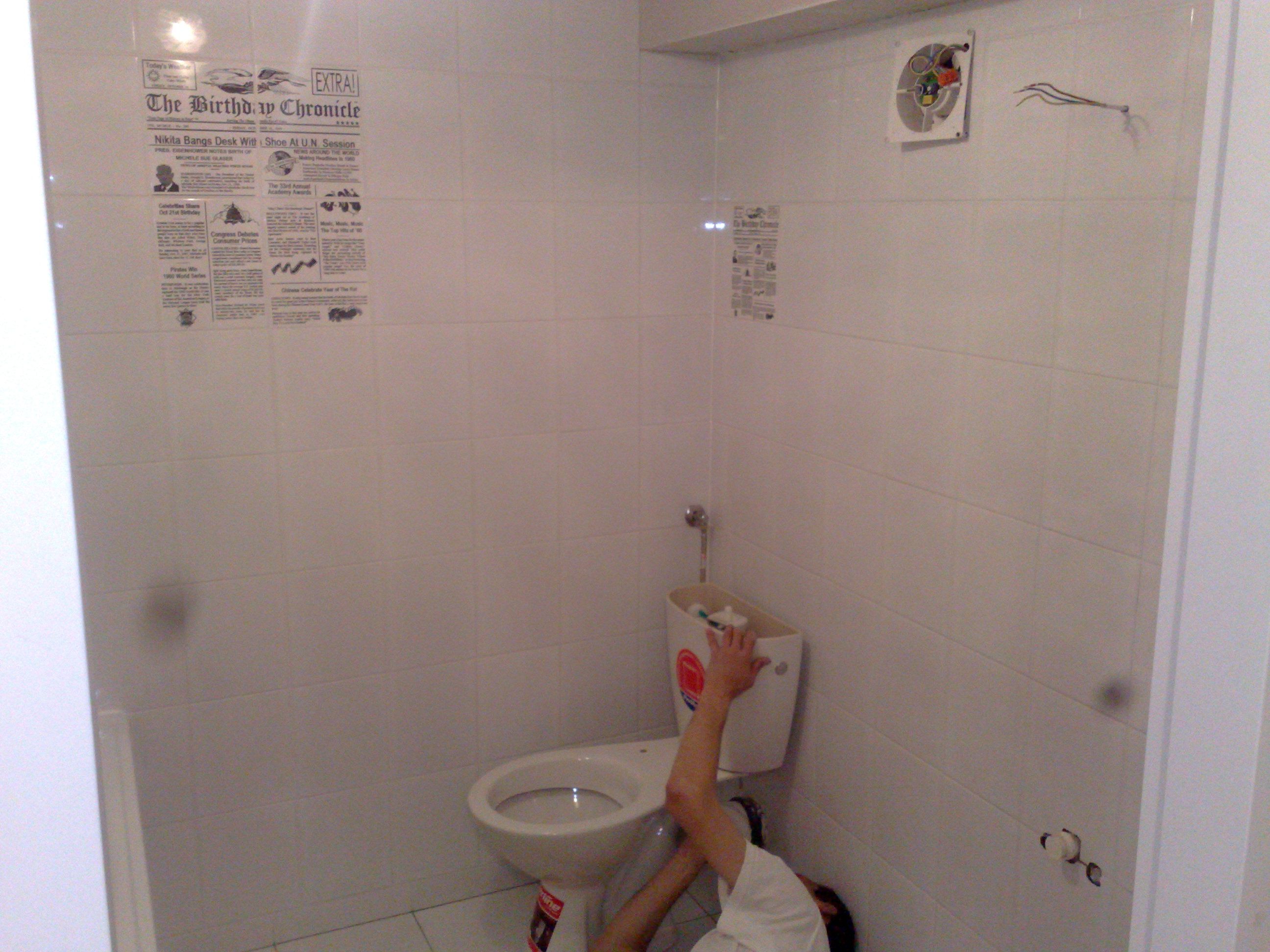 Montaż toalety w małej, wykafelkowanej łazience z gazetą przyklejoną do ściany, widoczny fragment osoby pracującej nad instalacją.