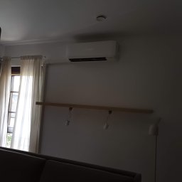 Frost Me - Biały klimatyzator Mitsubishi zamontowany na ścianie w pokoju z oknem zasłoniętym kremowymi zasłonami, widoczny fragment sofy i lampy stojącej.