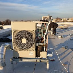 Frost Me - Montaż zewnętrznej jednostki klimatyzacyjnej na dachu budynku, widoczne otwarte panele, instalator w czarnym kombinezonie pochylony nad urządzeniem, przewody, narzędzia, w tle zabudowa miejska...