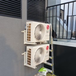 Frost Me - Dwa klimatyzatory LG Dual Inverter zamontowane na szarej ścianie budynku z widoczną kratką wentylacyjną, rurami wentylacyjnymi, drabiną i workiem nawozu u podstawy ściany.