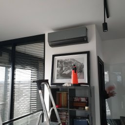 Frost Me - Szary klimatyzator LG zamontowany na ścianie w nowoczesnym wnętrzu, widoczna drabina przystawiona do regału i osoba przechodząca przez szklane drzwi.