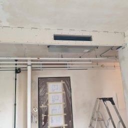 Frost Me - Montaż kanałów wentylacyjnych i klimatyzacyjnych w surowym wnętrzu biura, widoczne przewody, drabina i zabezpieczone folią drzwi z planami technicznymi.