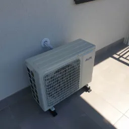 Zewnętrzna jednostka klimatyzacji AUX zamontowana na szarym tarasie, widoczna rurka w białej osłonie wchodząca w ścianę budynku, cień na płytkach.