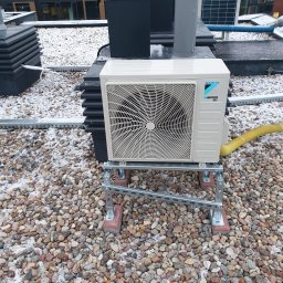 Frost Me - Biała jednostka zewnętrzna Daikin zamontowana na metalowej konstrukcji na dachu pokrytym żwirem z resztkami śniegu, widoczne rury wentylacyjne i budynek biurowy w tle.