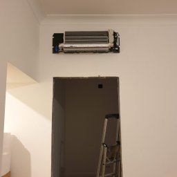 Frost Me - Demontaż klimatyzacji na białej ścianie z widoczną drabiną i otwartym przejściem do innego pomieszczenia.