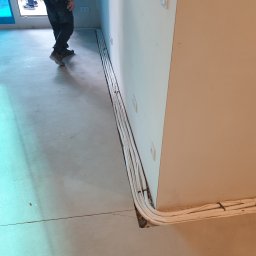 Frost Me - Montaż instalacji elektrycznej w nowym budynku w Warszawie, widoczne kable prowadzone wzdłuż ściany, częściowo ukryte w bruździe w podłodze. Osoba w tle w kratkowanej koszuli.