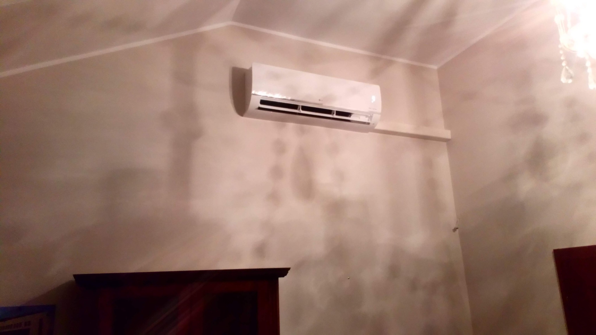 Biały klimatyzator zamontowany na ścianie w pokoju z dekoracyjnym wykończeniem, widoczny fragment szafki z ciemnego drewna w dolnej części kadru, oświetlenie górne z elementami kryształowymi.