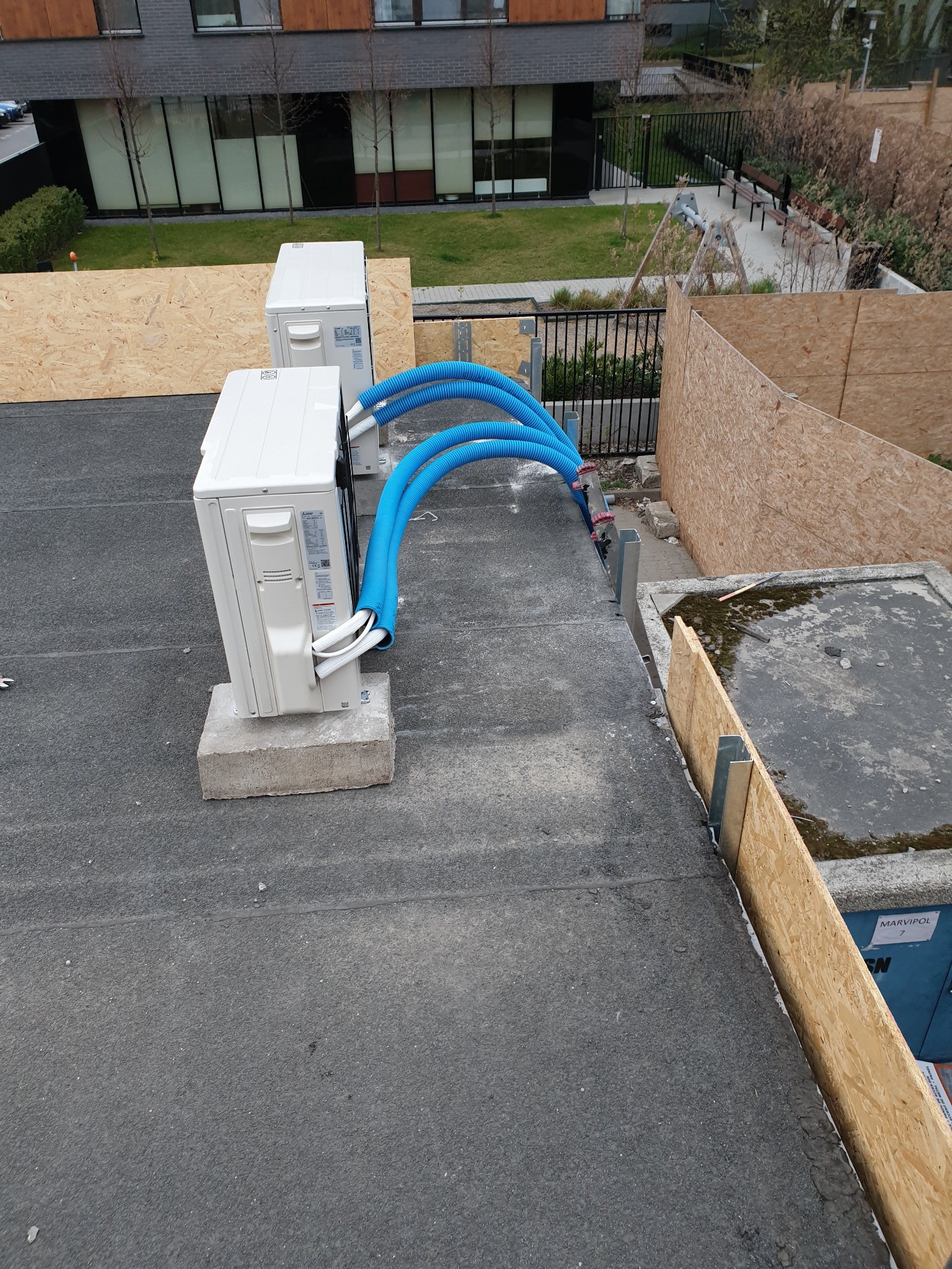 Dwa białe agregaty klimatyzacyjne na betonowych podstawach, połączone niebieskimi rurami, zamontowane na płaskim dachu budynku, widoczne elementy instalacji i otoczenie budynku.