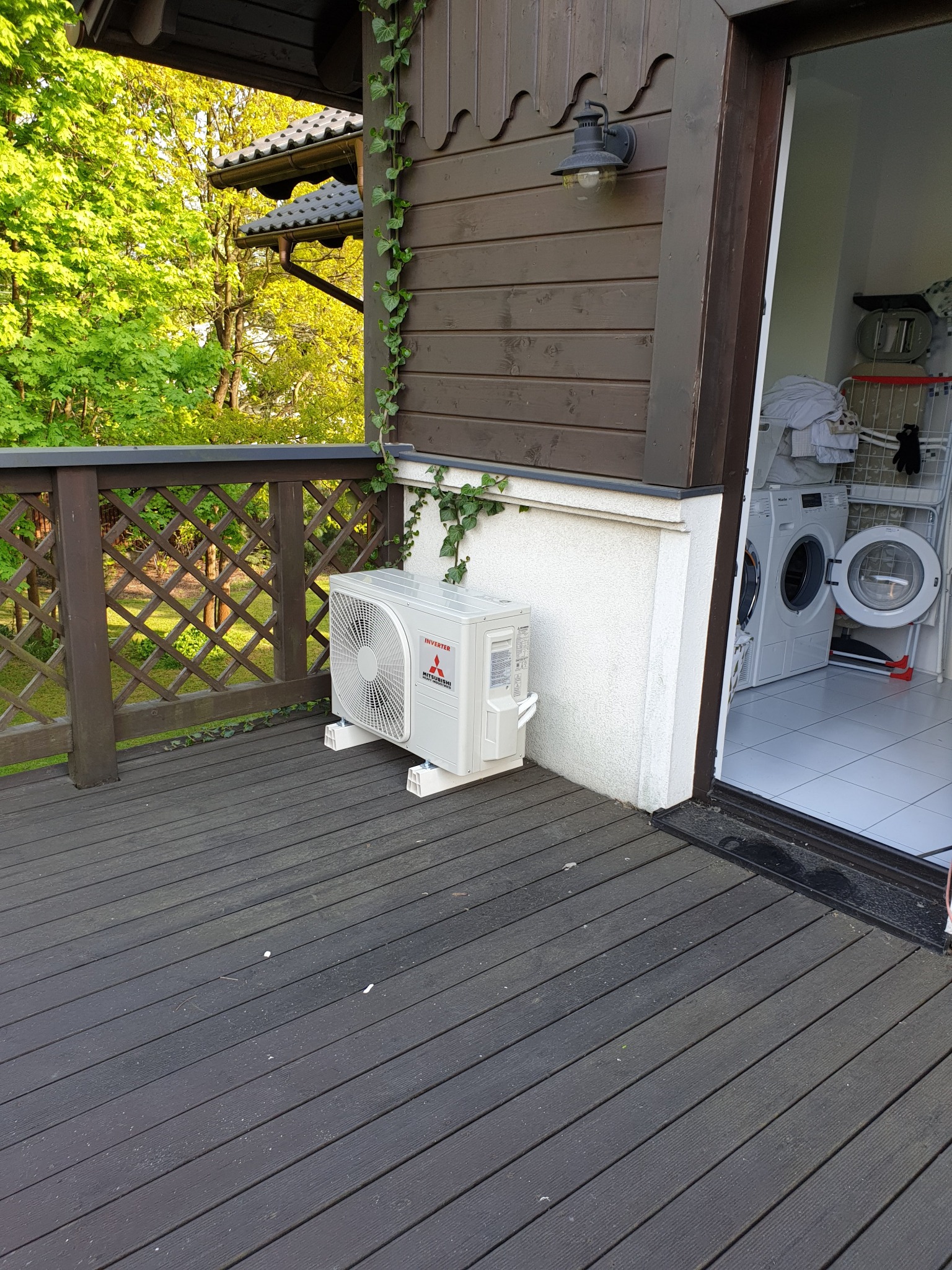 Zewnętrzna jednostka klimatyzacji Mitsubishi Electric zamontowana na drewnianym tarasie przy ścianie budynku z dekoracyjnym pnączem, widok na pralnię z pralkami.