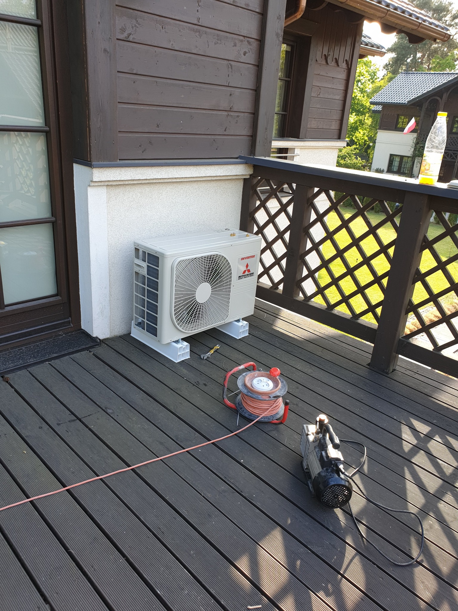 Zewnętrzna jednostka klimatyzatora Mitsubishi Inverter zamontowana na tarasie drewnianego domu, obok narzędzia serwisowe: pompa próżniowa i przedłużacz na bębnie.