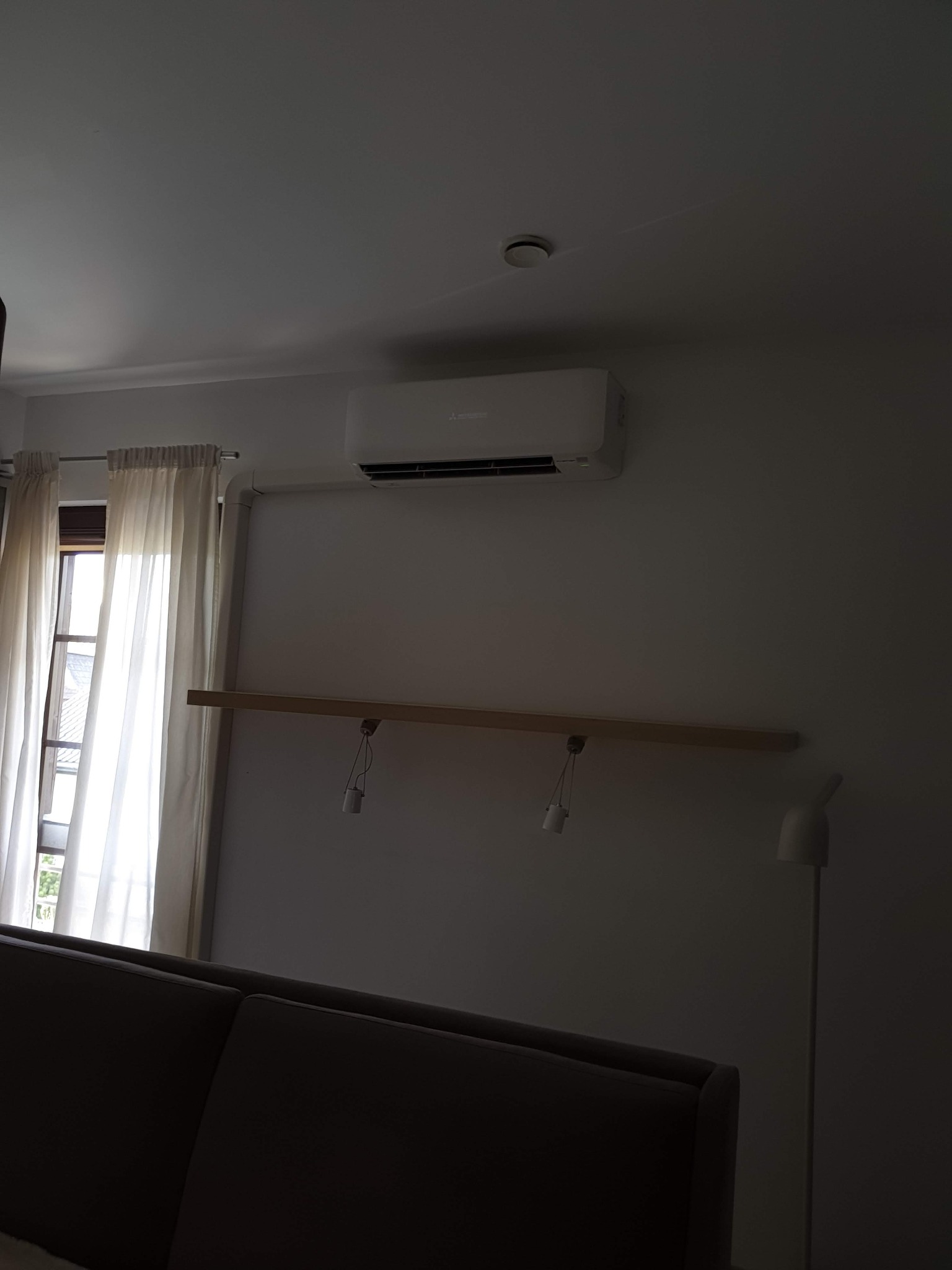 Biały klimatyzator Mitsubishi zamontowany na ścianie w pokoju z oknem zasłoniętym kremowymi zasłonami, widoczny fragment sofy i lampy stojącej.