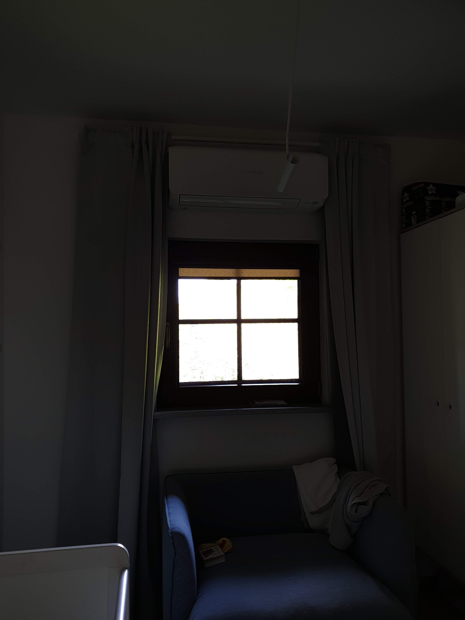 Wnętrze pokoju z zainstalowanym klimatyzatorem nad oknem, zasłonięte szare zasłony, niebieski fotel z narzuconym ręcznikiem.