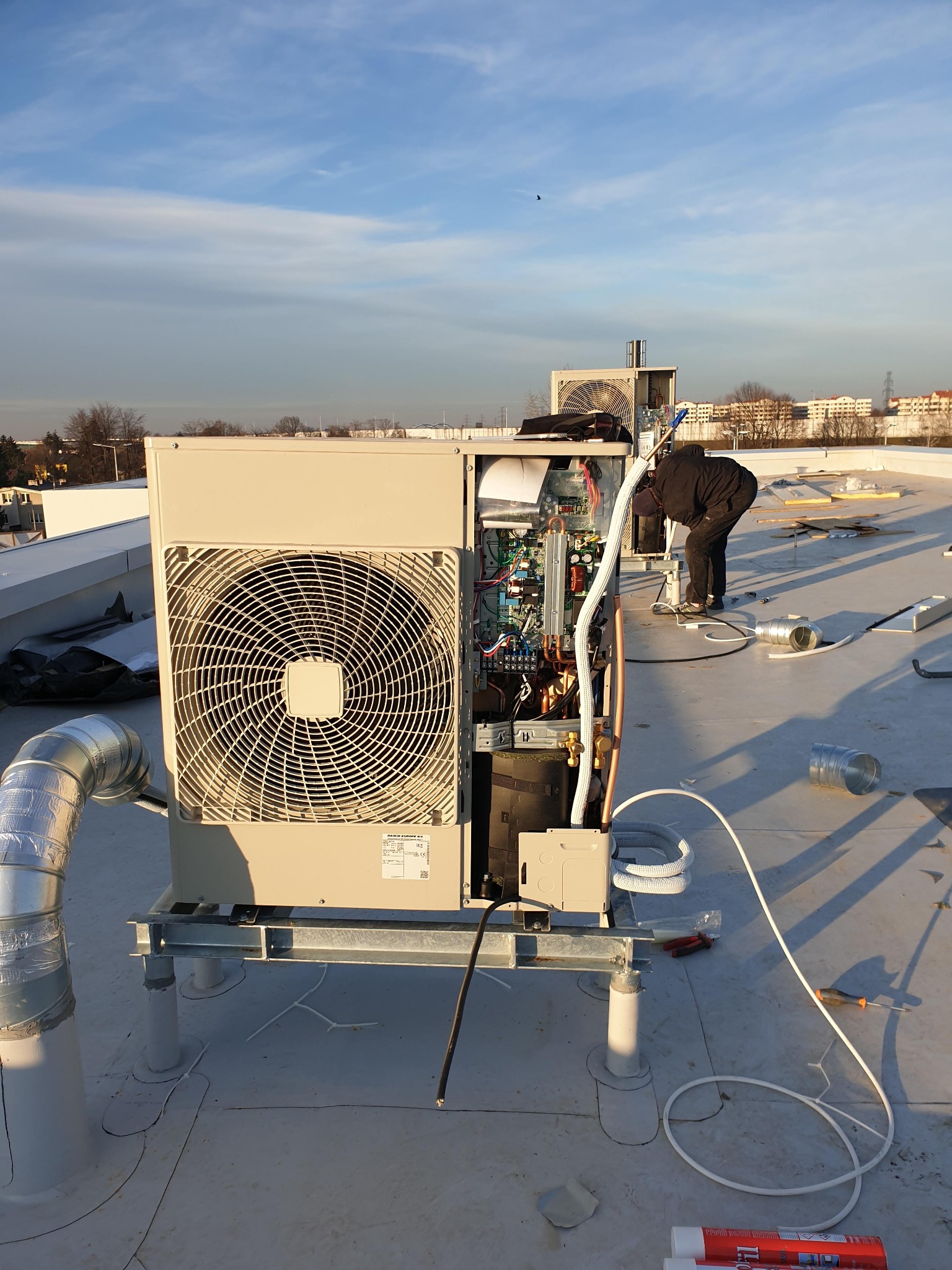 Montaż zewnętrznej jednostki klimatyzacyjnej na dachu budynku, widoczne otwarte panele, instalator w czarnym kombinezonie pochylony nad urządzeniem, przewody, narzędzia, w tle zabudowa miejska...