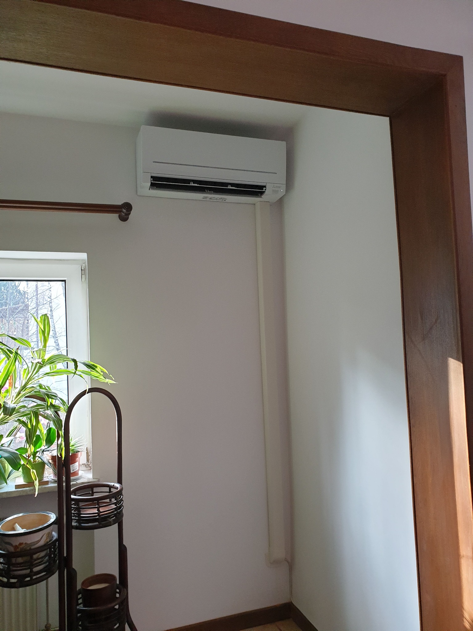 Biały klimatyzator ścienny zamontowany w rogu pokoju, widoczny zza drewnianej futryny drzwiowej, obok okna z roślinami doniczkowymi i metalowym kwietnikiem.