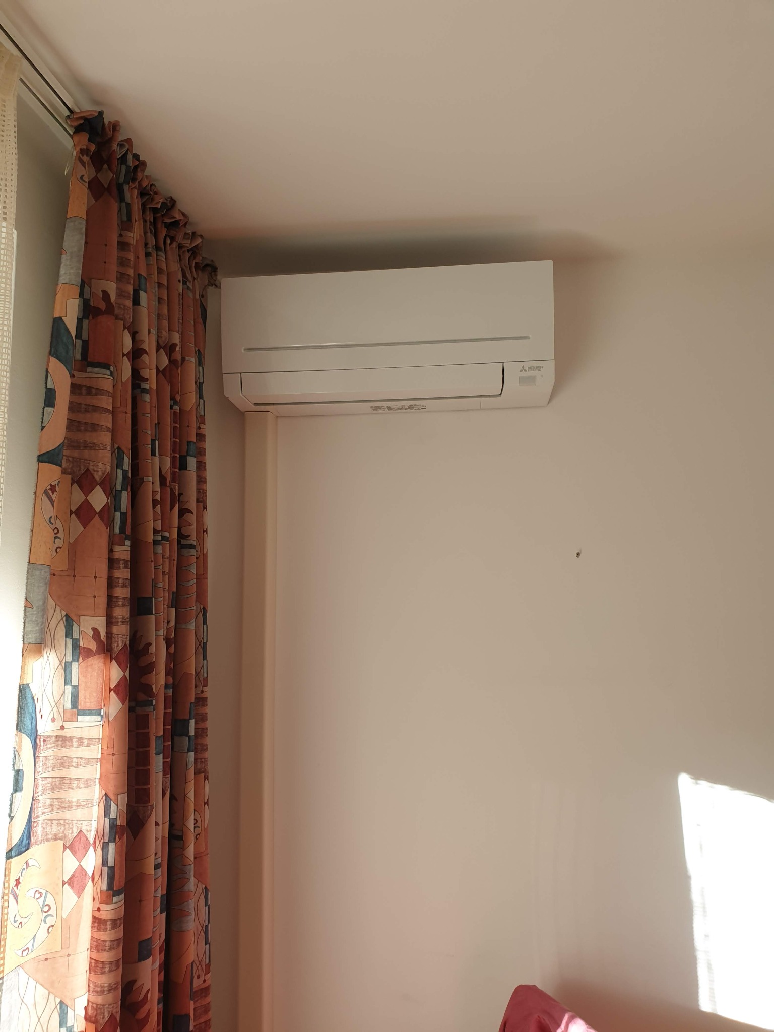Biały klimatyzator Mitsubishi Electric zamontowany na jasnej ścianie w pokoju, obok wzorzystych zasłon w odcieniach brązu i beżu.