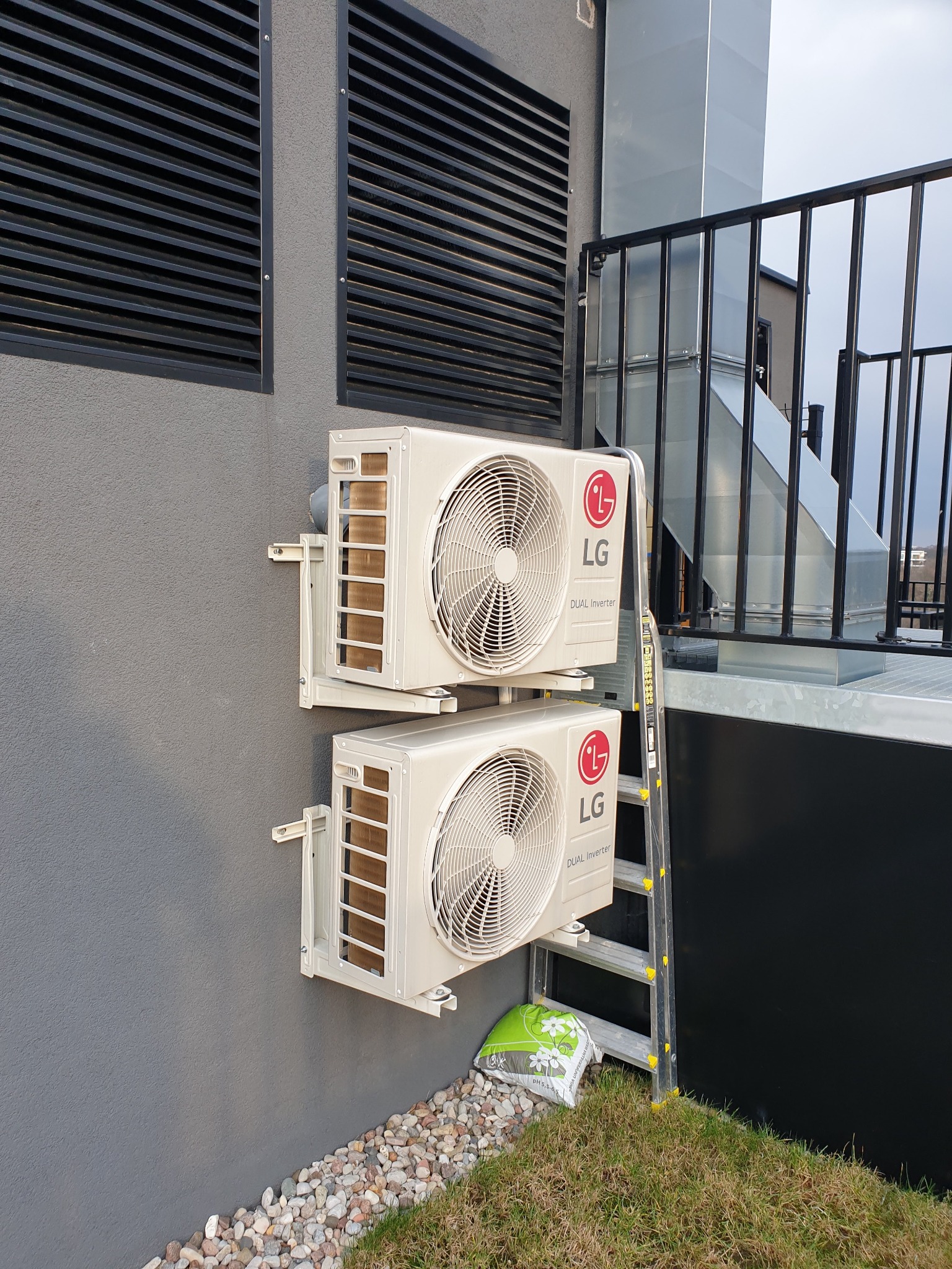 Dwa klimatyzatory LG Dual Inverter zamontowane na szarej ścianie budynku z widoczną kratką wentylacyjną, rurami wentylacyjnymi, drabiną i workiem nawozu u podstawy ściany.