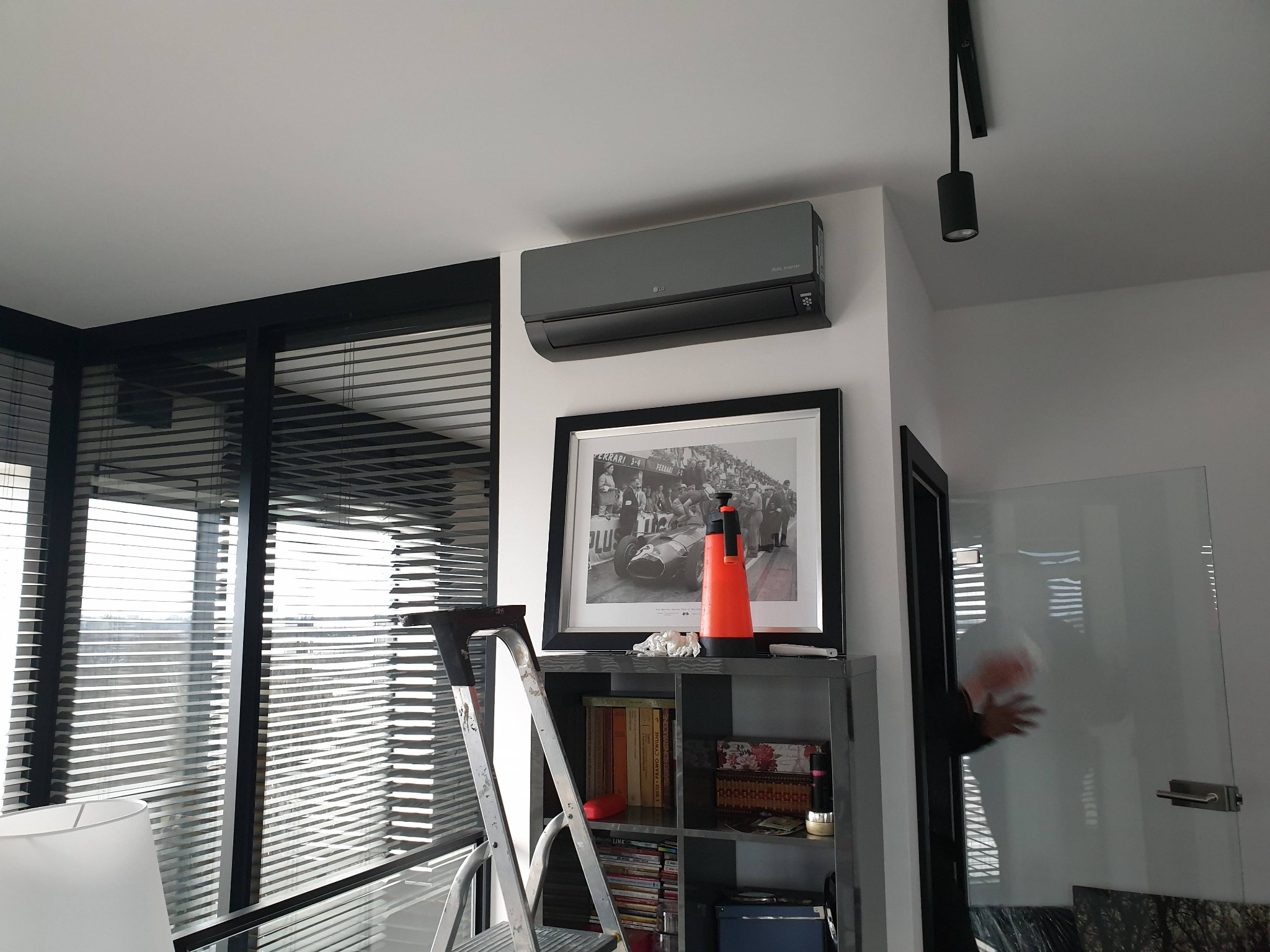 Szary klimatyzator LG zamontowany na ścianie w nowoczesnym wnętrzu, widoczna drabina przystawiona do regału i osoba przechodząca przez szklane drzwi.