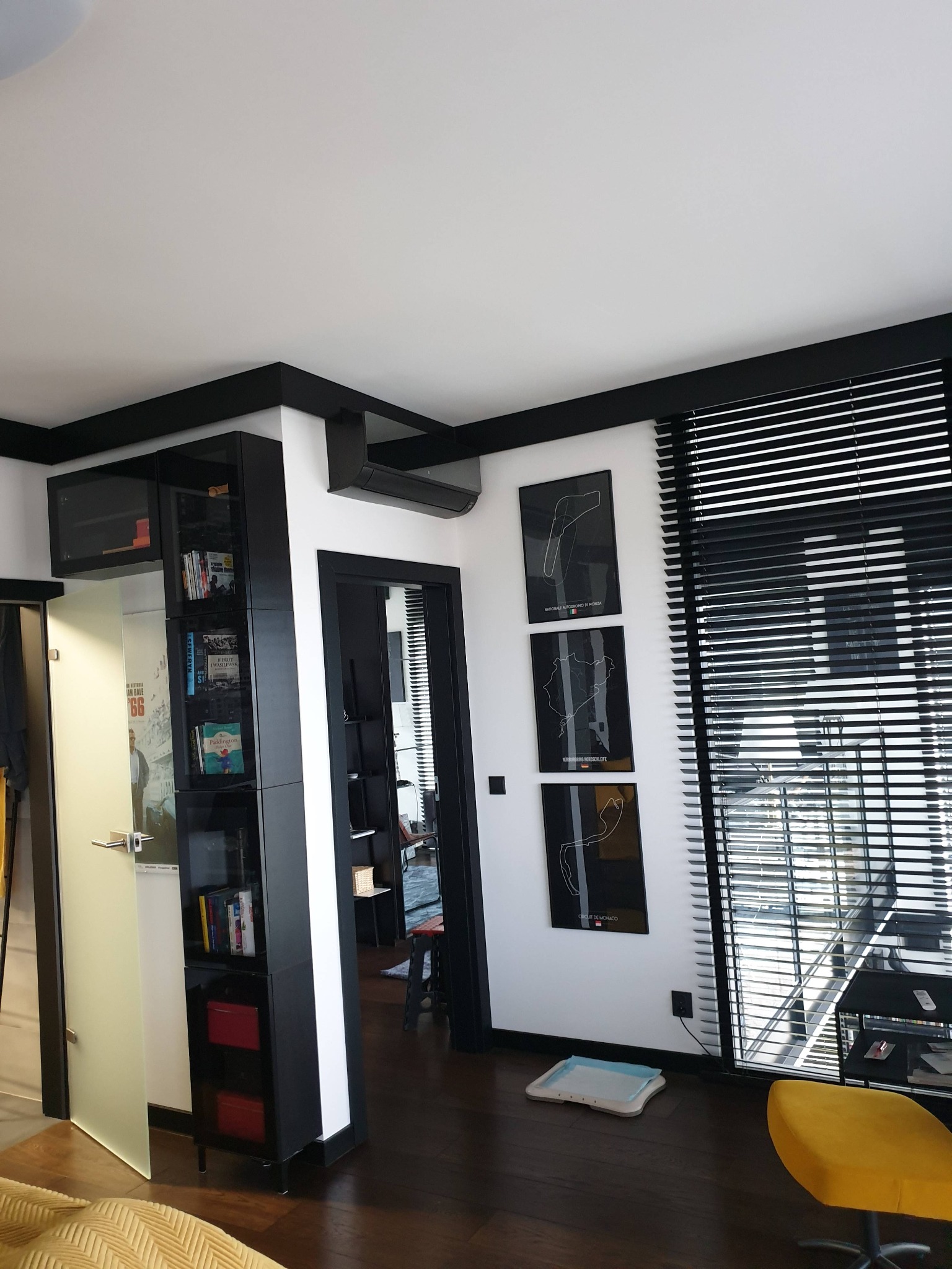 Wnętrze pokoju z zainstalowanym klimatyzatorem ściennym, widoczne czarne żaluzje, minimalistyczne plakaty z torami wyścigowymi i czarne meble