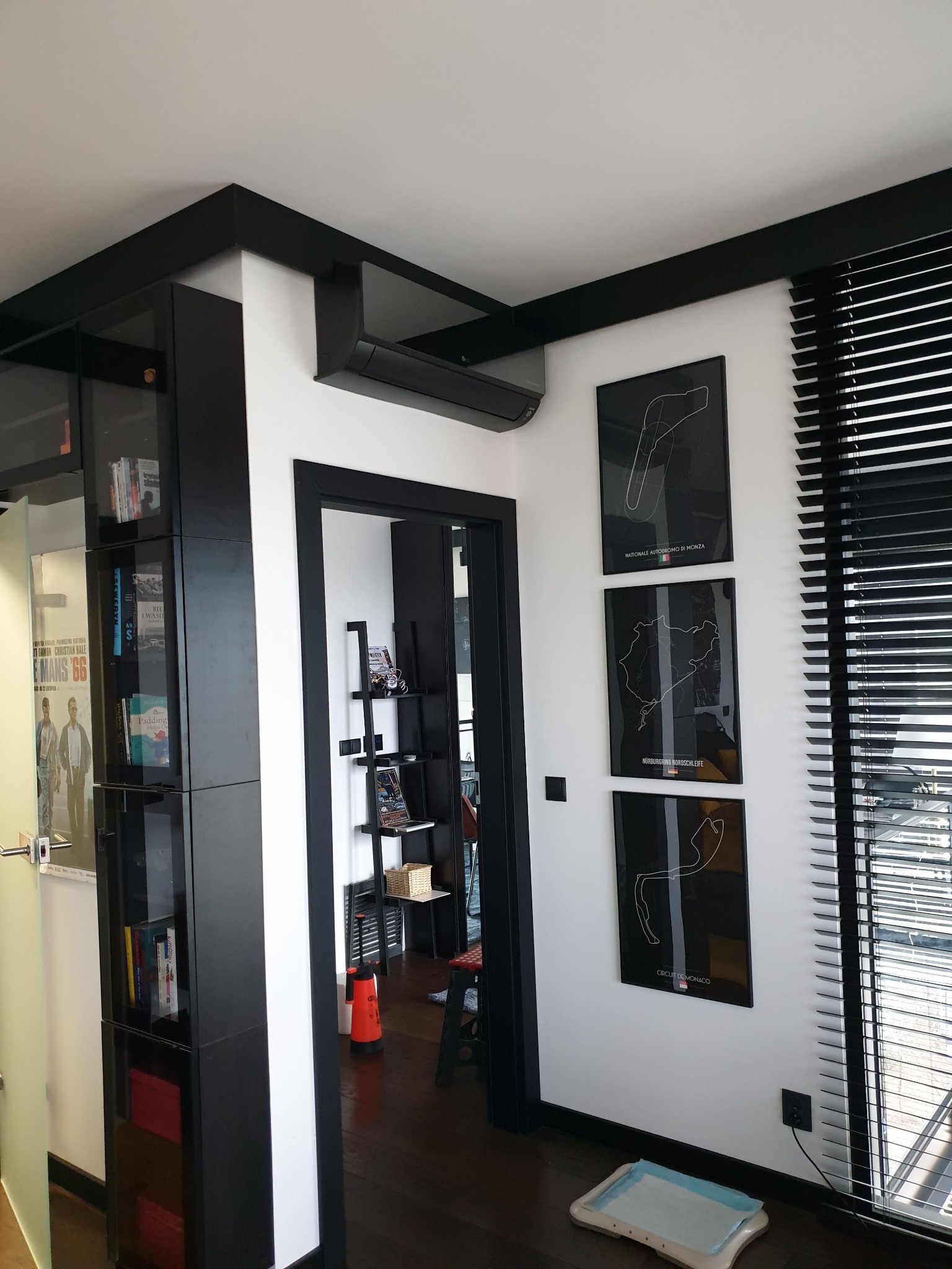 Wnętrze z zamontowaną jednostką klimatyzacyjną na ścianie nad drzwiami, czarne ramy drzwi i mebli, plakaty z torami wyścigowymi na ścianie, żaluzje okienne.
