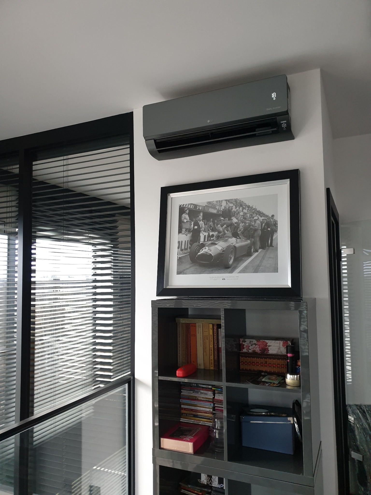 Szara jednostka wewnętrzna klimatyzatora LG z wyświetlaczem temperatury, zamontowana nad czarno-białym zdjęciem w ramce, umieszczonym na szarym regale z książkami i dekoracjami w mieszkaniu...