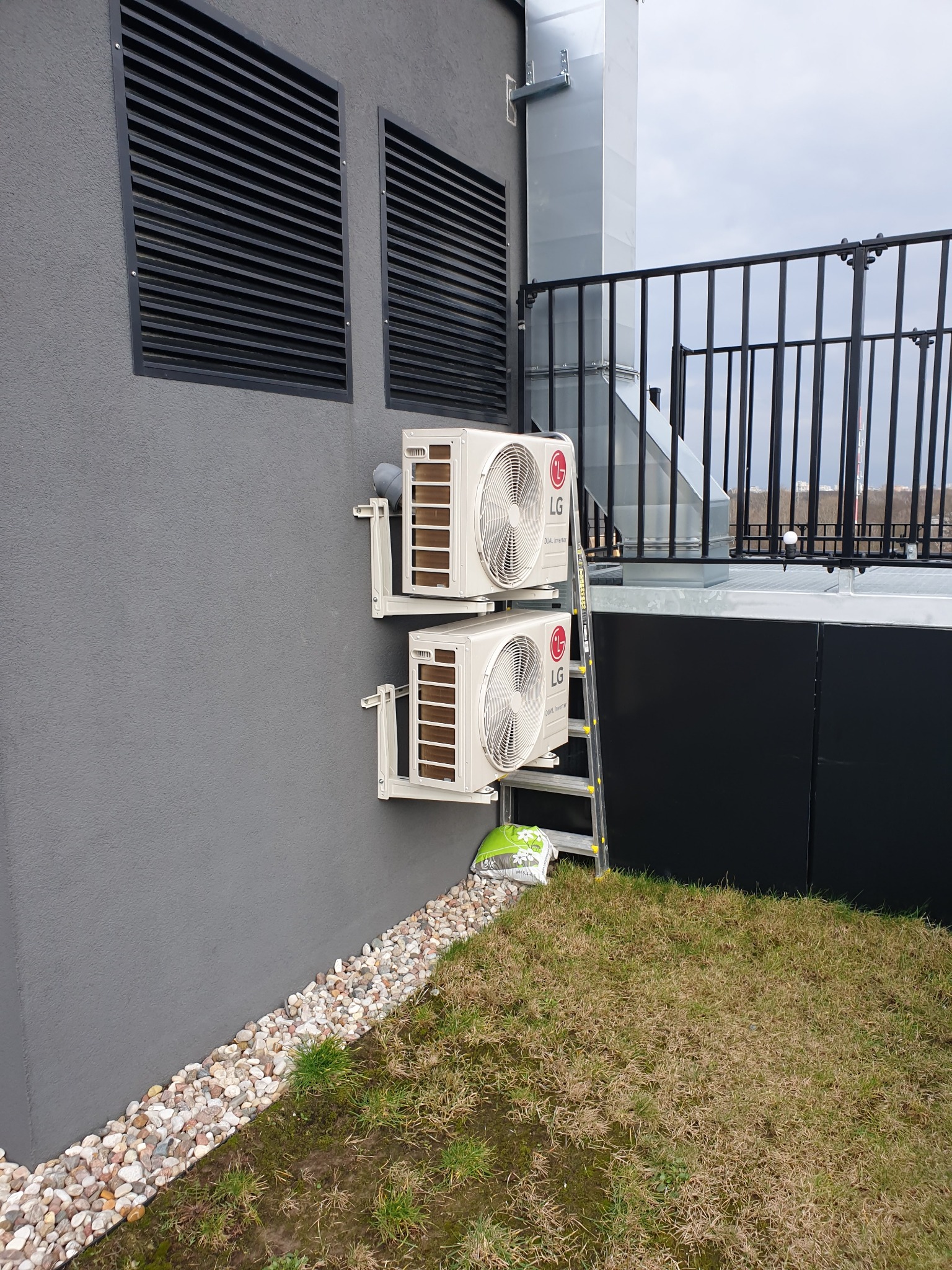 Dwa zewnętrzne moduły klimatyzacyjne LG zamontowane na szarej ścianie budynku, z widoczną drabiną opartą o moduły, obok leży worek z ziemią, na dachu widoczne barierki i komin wentylacyjny.