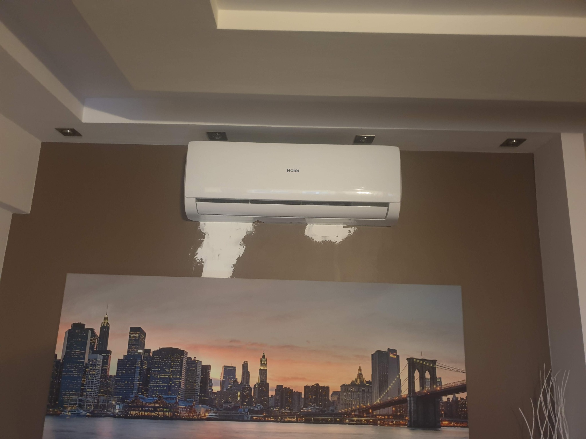 Biały klimatyzator Haier zamontowany na brązowej ścianie nad obrazem panoramy Nowego Jorku, ślady po szpachlowaniu pod klimatyzatorem.