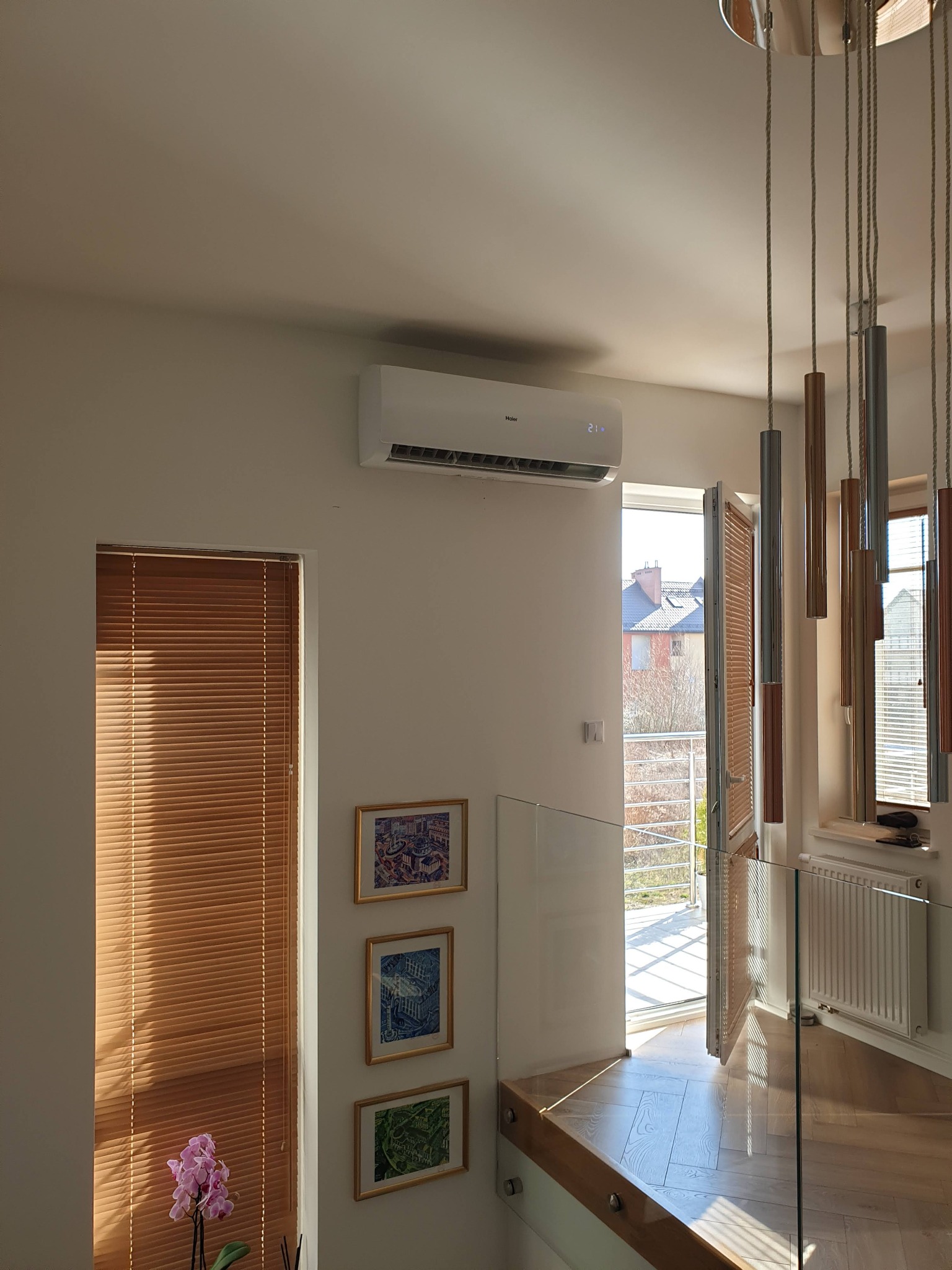 Nowoczesny klimatyzator Haier zamontowany na ścianie w jasnym wnętrzu z drewnianymi żaluzjami i trzema obrazkami w złotych ramkach, widok z góry na balustradę schodów i otwarte drzwi balkonowe.