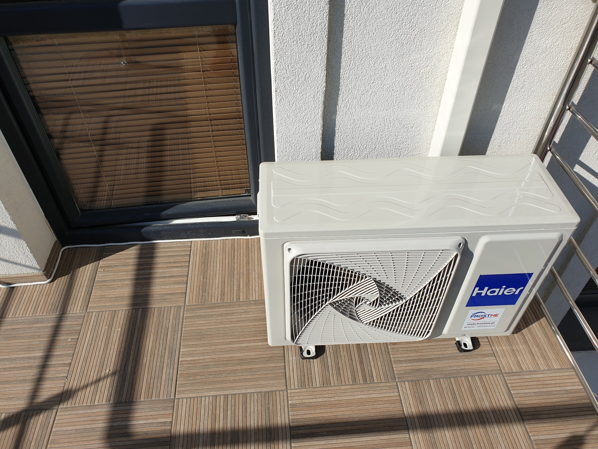 Zewnętrzna jednostka klimatyzatora Haier zamontowana na balkonie z brązowymi płytkami, widoczne rolety w oknie i fragment balustrady.