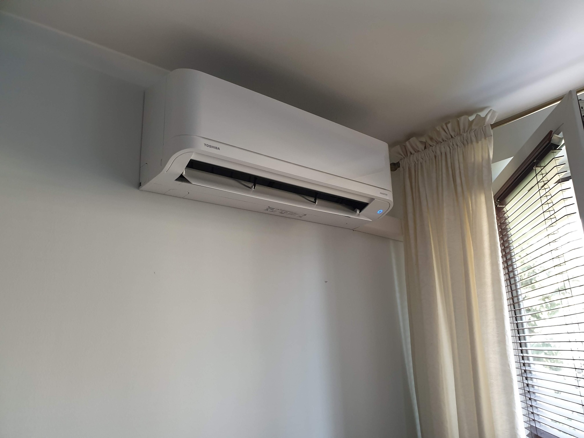 Biała jednostka wewnętrzna klimatyzatora Toshiba zamontowana na ścianie w jasnym pomieszczeniu, z widocznym oknem z żaluzjami i kremowymi zasłonami.
