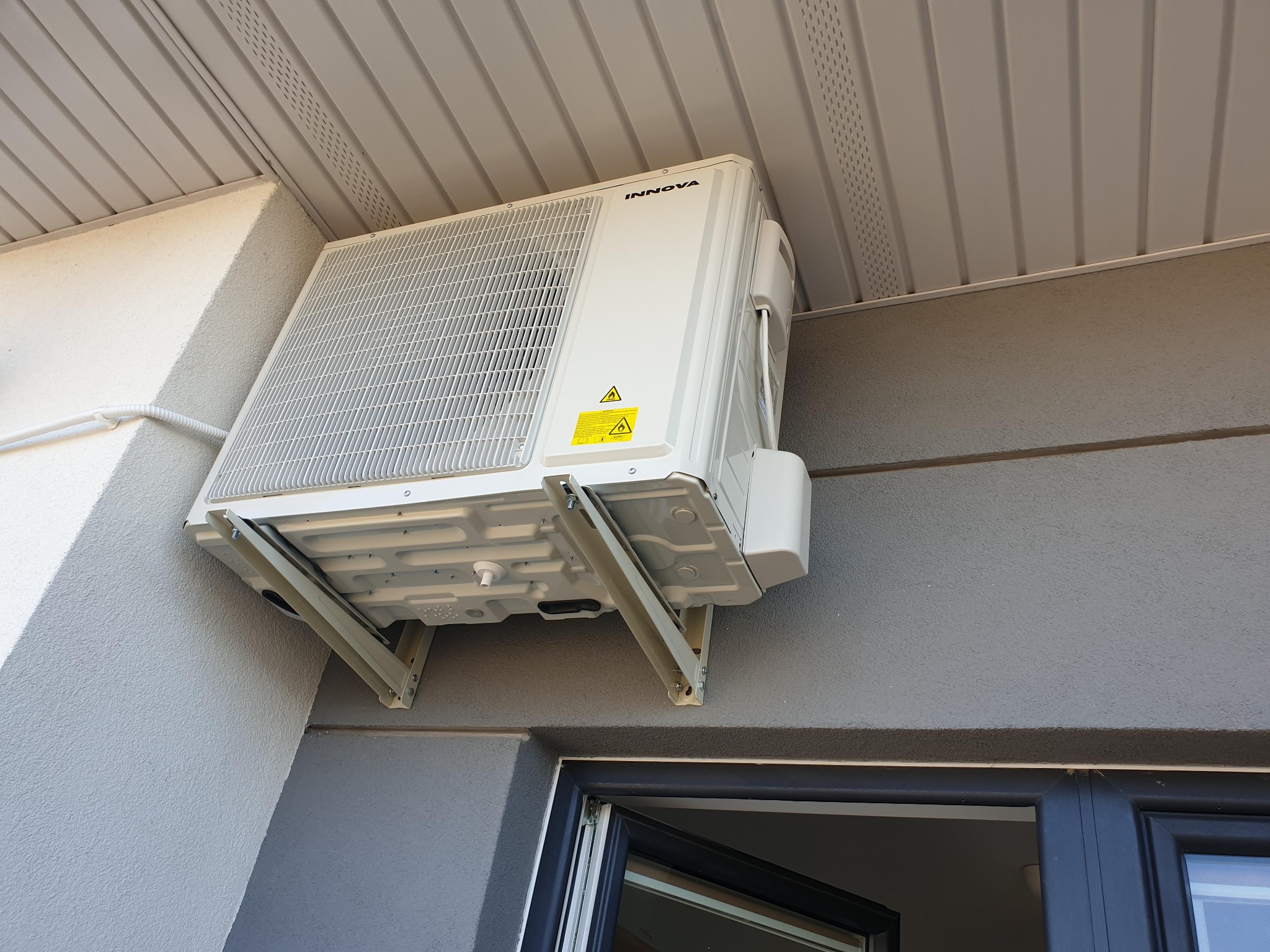Biała jednostka zewnętrzna marki Innova zamontowana na szarej elewacji budynku z widocznymi wspornikami montażowymi i fragmentem okna.