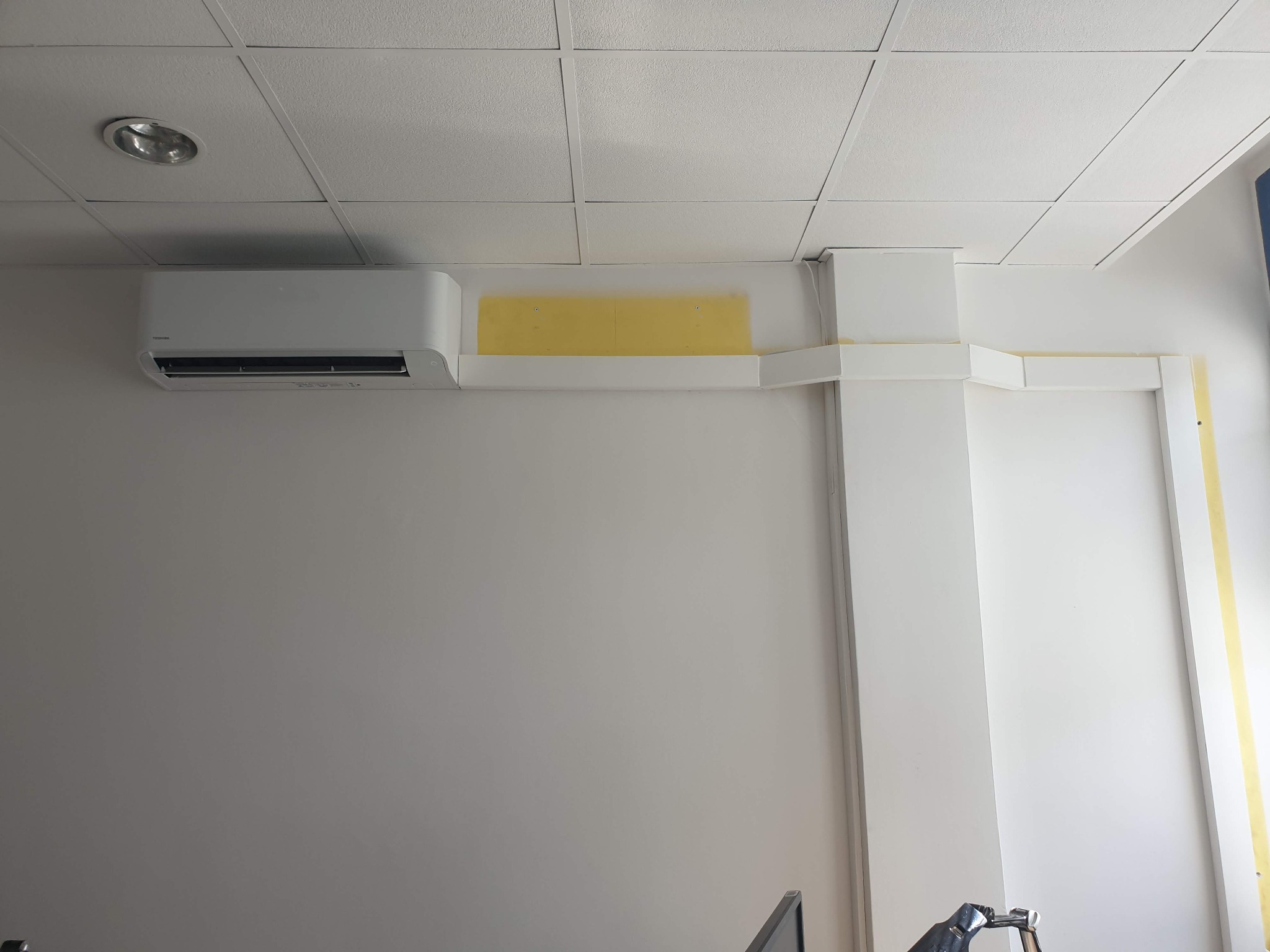 Biała klimatyzacja Toshiba zamontowana na ścianie w biurze, widoczne żółte taśmy zabezpieczające podczas montażu, kratka sufitowa w tle.