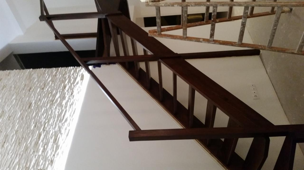 Ciemnobrązowe drewniane schody z balustradą, widoczne z dołu, z fragmentem ściany pokrytej dekoracyjnym kamieniem i drabina malarska w tle.