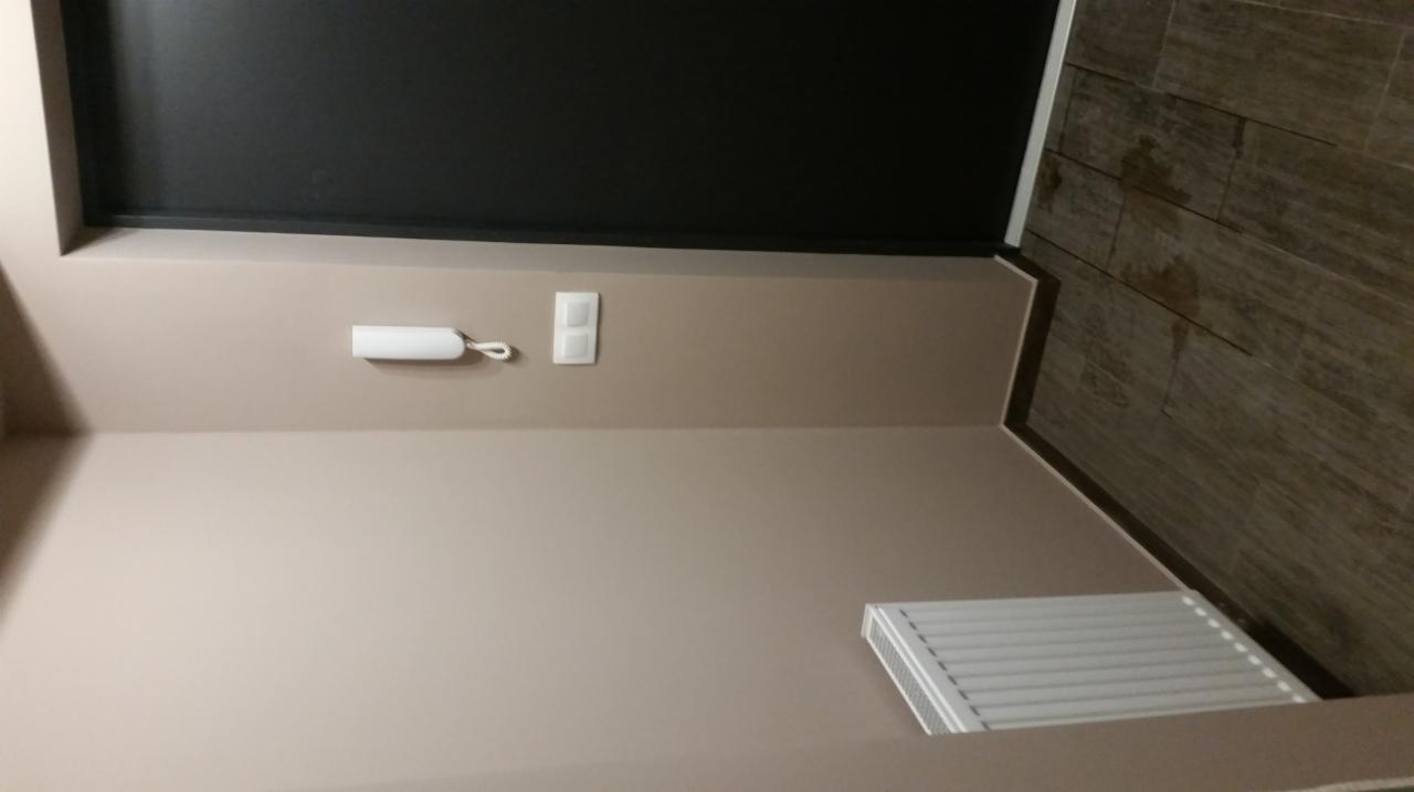 Pokój po malowaniu ścian na beżowo, widoczny grzejnik, domofon i włącznik światła, oraz fragment ściany wyłożonej płytkami imitującymi drewno.