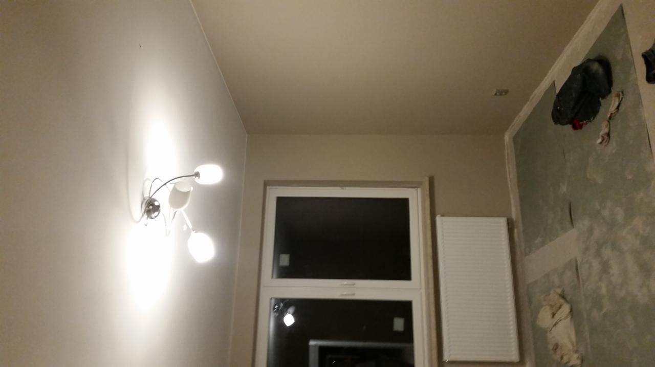 Ściana w trakcie przygotowania do malowania, widoczne ślady po szpachlowaniu i zabezpieczenia folią, nowoczesna lampa ścienna z trzema kloszami, okno z białymi ramami.