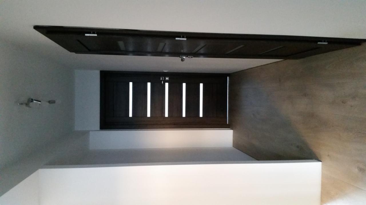 Ciemne, drewniane drzwi wewnętrzne z przeszkleniami w postaci pionowych pasków, zamontowane w białej ścianie, widoczne z perspektywy podłogi, na tle ściany wykończonej panelami drewnopodobnymi.