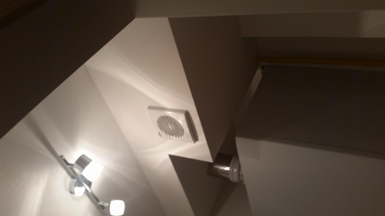 Biały wentylator łazienkowy zamontowany na suficie pod skosem, oświetlony przez chromowaną lampę z dwoma punktami świetlnymi, widoczne elementy instalacji wentylacyjnej.
