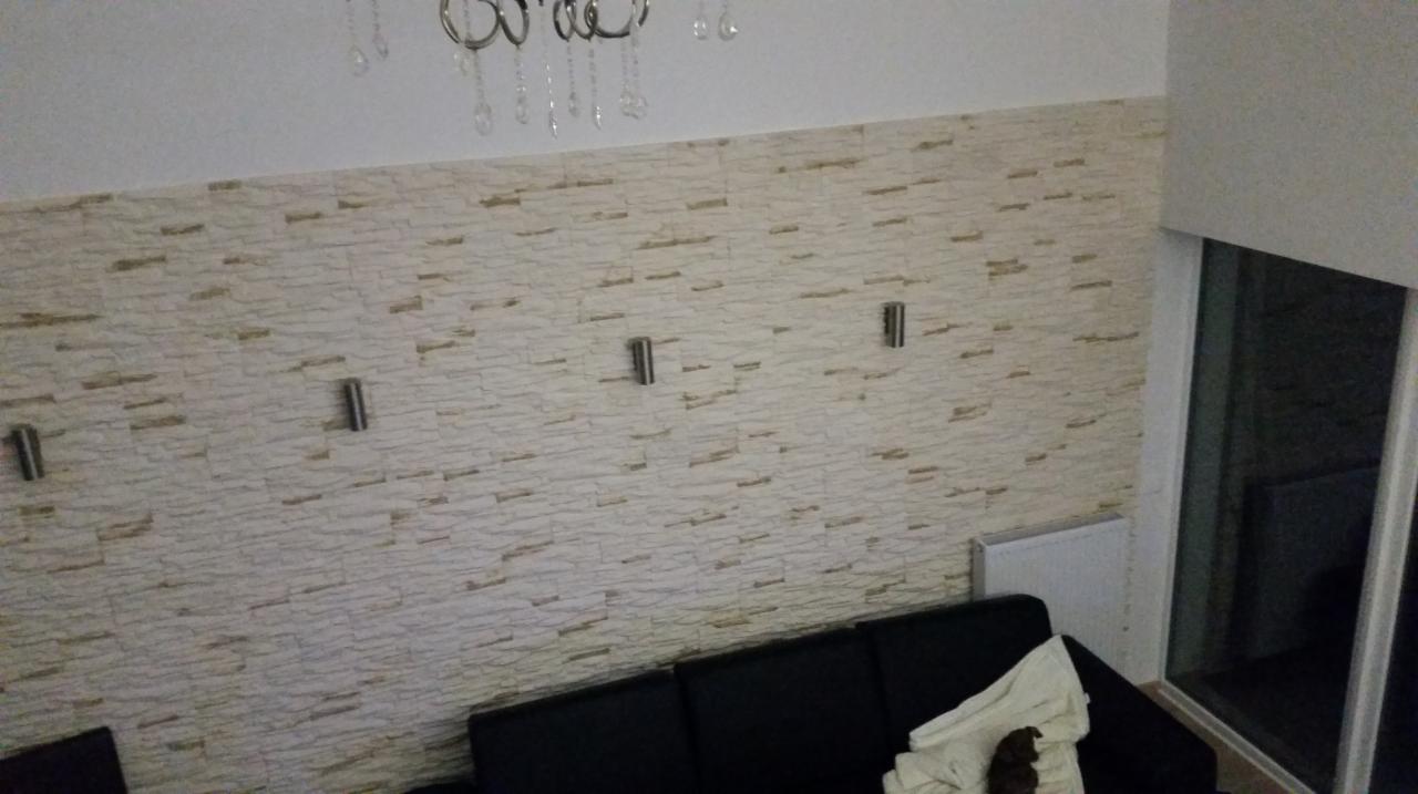 Ściana wykończona tapetą imitującą kamień z zamontowanymi metalowymi kinkietami, widoczny fragment czarnej sofy z jasnym pledem i małym psem.