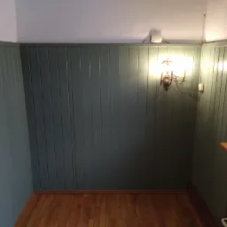 Pomieszczenie z panelami ściennymi w odcieniach zieleni, drewniana podłoga, kinkiet z żarówkami imitującymi świece i widoczne elementy instalacji elektrycznej pod sufitem.