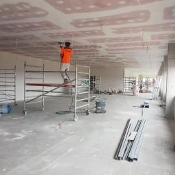 DeWall - Pracownik w pomarańczowej koszulce szlifuje sufit z płyt gipsowo-kartonowych na rusztowaniu w surowym wnętrzu biurowym przed malowaniem, widoczne narzędzia i materiały budowlane.