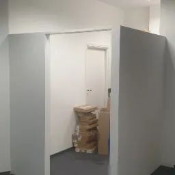 Wnętrze biura po malowaniu ścian na biało, z widocznymi kartonami w rogu pomieszczenia i szarą wykładziną na podłodze.