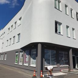 DeWall - Malowanie elewacji biurowca na kolor szary, widoczny malarz przy pracy, zabezpieczone okna folią, pachołki ostrzegawcze, błękitne niebo w tle.