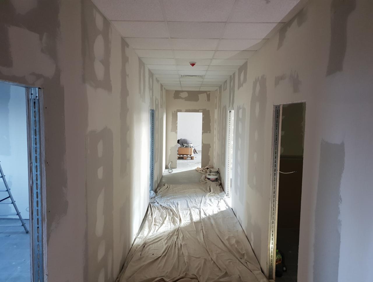 Korytarz biurowy w trakcie szpachlowania ścian, widoczne zakryte podłogi, drabina i pracownik w oddali