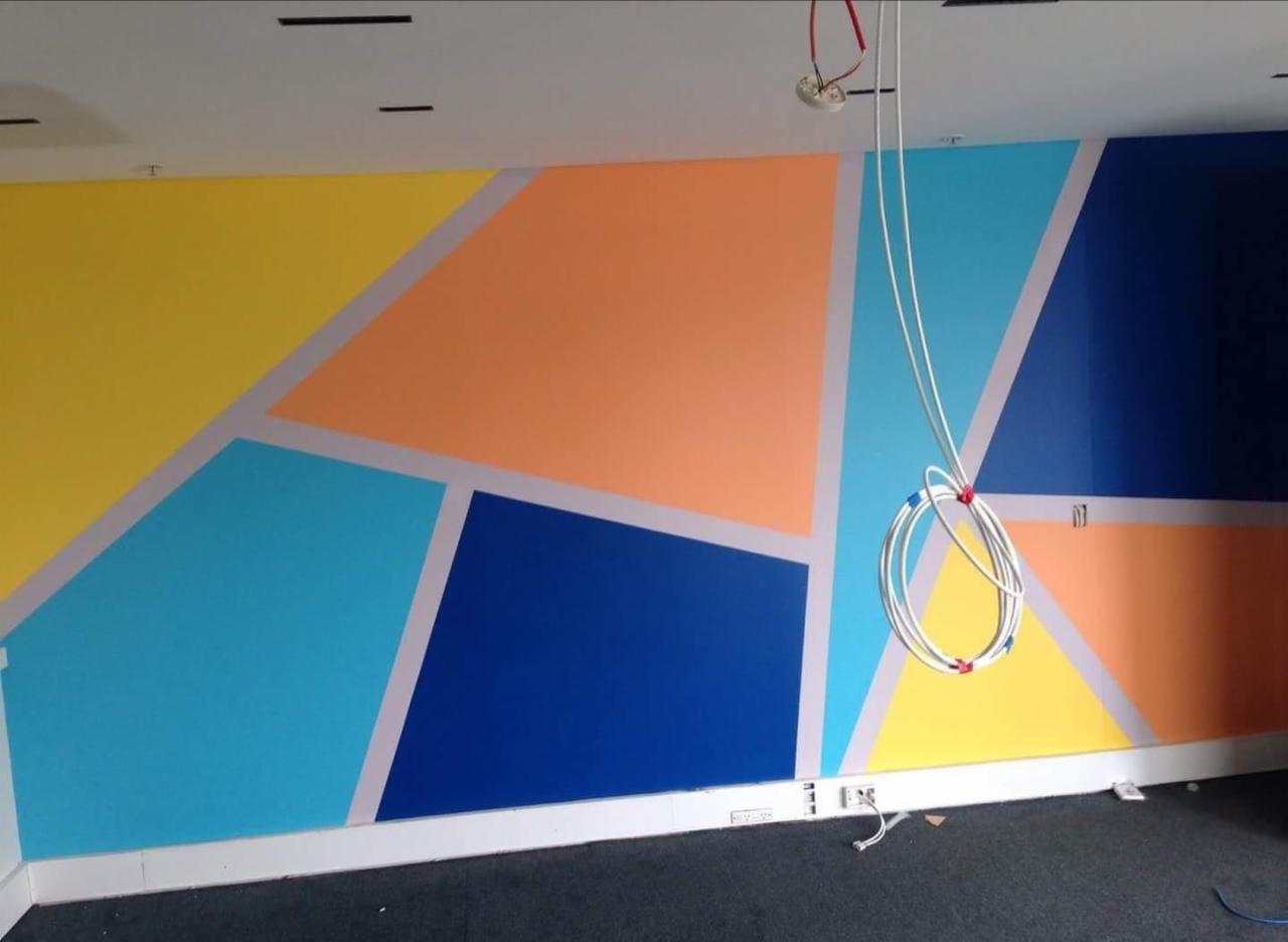 Ściana w biurze pomalowana w geometryczne wzory w kolorach żółtym, pomarańczowym, niebieskim i granatowym, oddzielone szarymi paskami. Widoczne wystające kable elektryczne i gniazdka w dolnej...