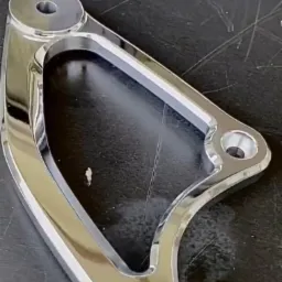 Błyszczący, chromowany element metalowy z otworami, leżący na ciemnej powierzchni. Widoczne refleksy światła podkreślają gładką powierzchnię.