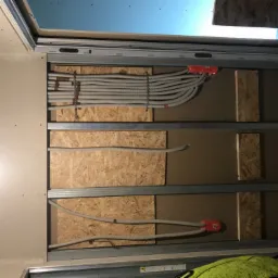 Wewnętrzna ściana w trakcie budowy z widocznymi rurami karbowanymi z przewodami, zamocowanymi do płyty OSB i metalowych profili, z widoczną puszką instalacyjną w kolorze czerwonym.