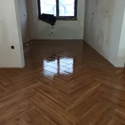 Ułożona drewnopodobna podłoga z płytek w jodełkę w trakcie remontu, widoczne ślady po pracach na ścianach, okno z widokiem na sąsiedni budynek.