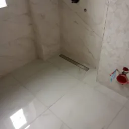 Wykończona kabina prysznicowa z odpływem liniowym, białe płytki imitujące marmur na ścianach i podłodze, bateria podtynkowa w kolorze czarnym, fragment ściany wyłożony mozaiką w geometryczny wzór.