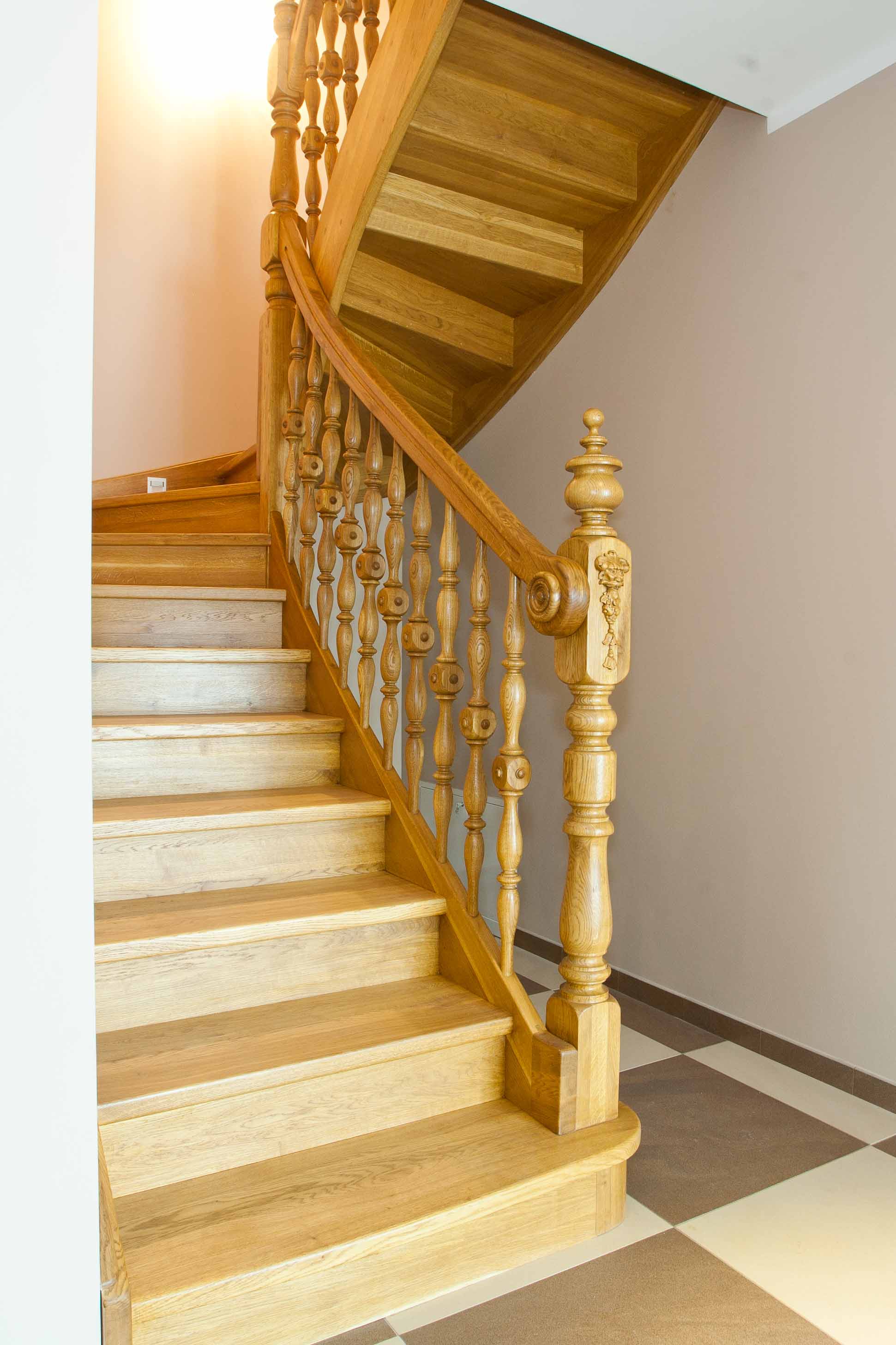 Jasne, drewniane schody z ozdobną balustradą z toczonymi elementami i rzeźbioną kolumną, widok z dołu, na podłodze płytki w szachownicę.