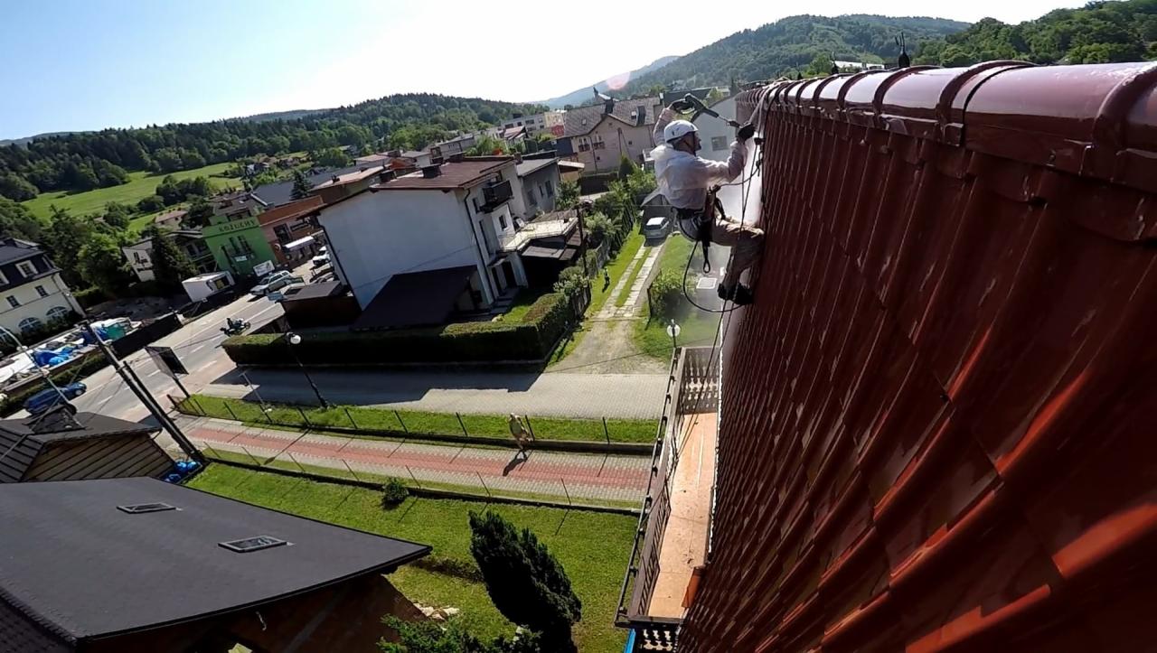 Pracownik w białym kombinezonie i kasku, zabezpieczony linami, czyści czerwoną dachówkę za pomocą myjki ciśnieniowej na tle zabudowy miejskiej i zielonych wzgórz.